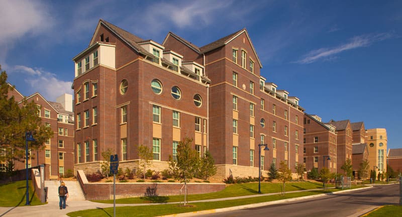 red brick residential hall building with 4 stories, blue sky in background and green grass on strip near blacktop of parking lot
